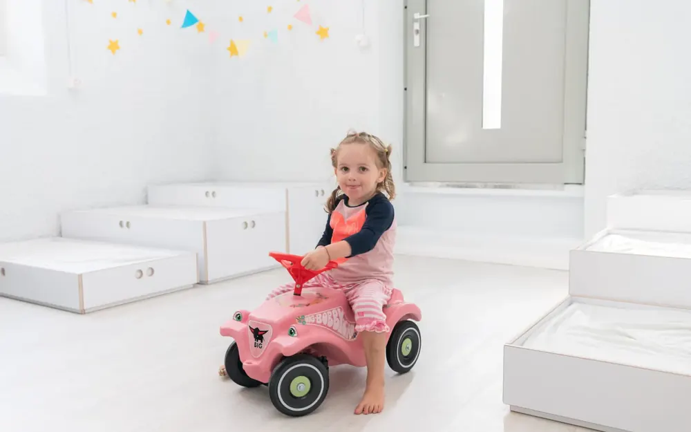 Kind fährt mit dem Rutschauto durch einen hellen Spielraum der Kita Bärlis